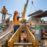 Construction workers wearing PPE addressing fall protection, crane lift, trench box inspection, and electrical safety on a daylight jobsite